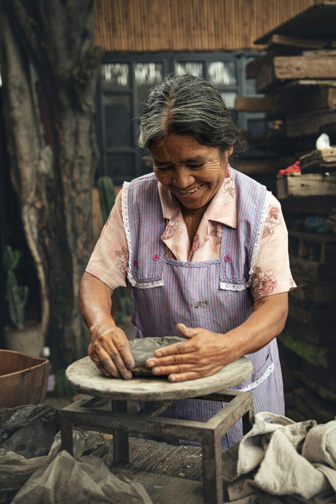 Elderly woman skillfully shaping clay by hand outdoors, showcasing traditional craftsmanship.